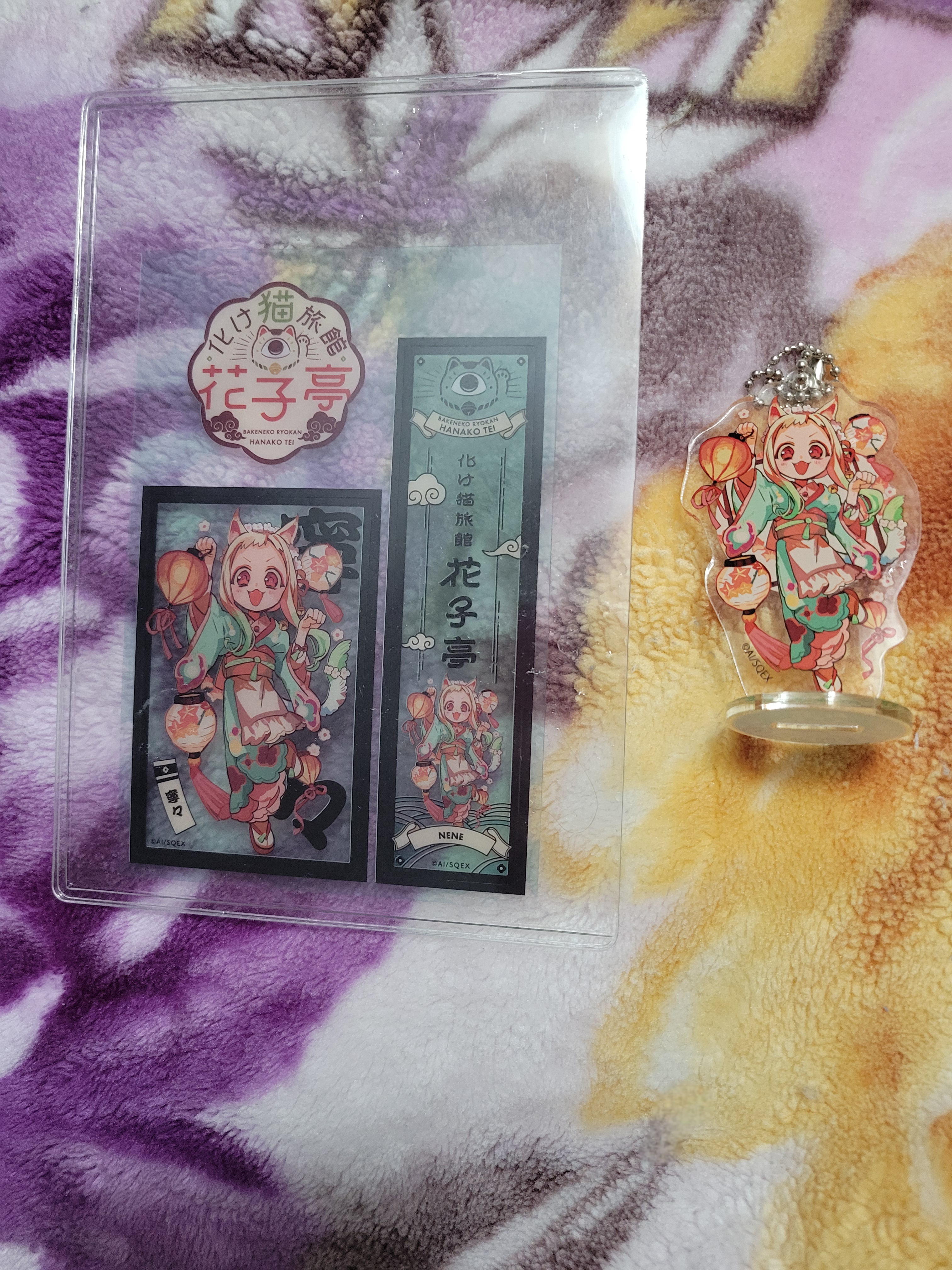 Acrylic standee, bookmark and print of Nene Yashiro in her Bakeneko Ryokan Hanakotei outfit. The art has her as a catgirl wearing a light blue kimono as she holds a lantern in one hand and the other is doing the cat paw. Lanterns float around her and there are flowers on her.